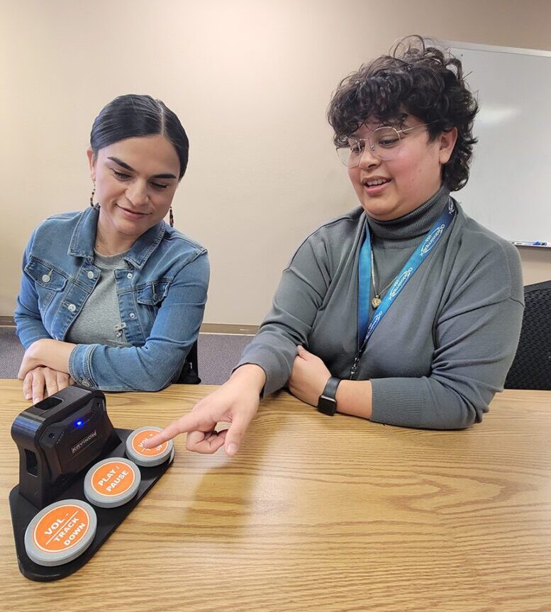 Pictured the AT Services Team Lizette and Michelle sitting at a table demonstrating a speaker adaptive device.