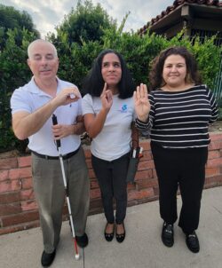 Pictured our Blind/Low-Vision Services team standing outside, smiling doing ASL Finger Spelling of O-I-B for our Older Individuals who are Blind – what our long-reputation is most known for. Left: Paul in a collared shirt, holding his mobility cane with his left hand as he makes the “o” with his right; Center: Danyelle with shoulder length dark hair making an ‘I’ in ASL with her right hand; and to the right is Wendy, wearing a black top with white stripes making the “b” in ASL with her right hand. The trio is standing in front of a brick planter with greenery behind them, blue sky and white clouds above.