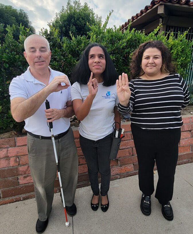 Pictured our Blind/Low-Vision Services team standing outside, smiling doing ASL Finger Spelling of O-I-B for our Older Individuals who are Blind – what our long-reputation is most known for.  Left: Paul in a collared shirt, holding his mobility cane with his left hand as he makes the “o” with his right;  Center: Danyelle with shoulder length dark hair making an ‘I’ in ASL with her right hand; and to the right is Wendy, wearing a black top with white stripes making the “b” in ASL with her right hand. The trio is standing in front of a brick planter with greenery behind them, blue sky and white clouds above.