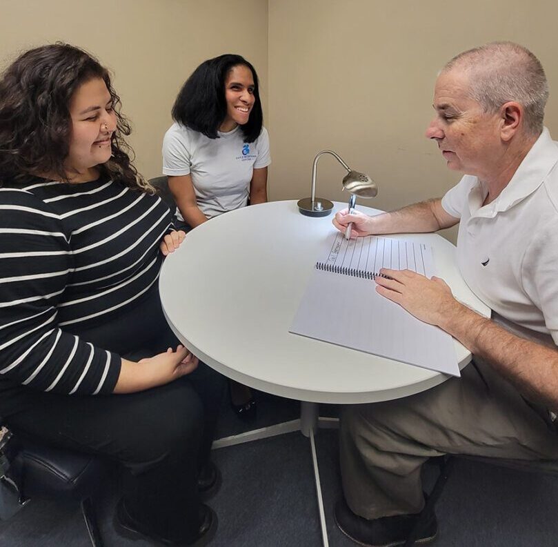 Sitting at a round table are DMC BLVS/Low-Vision Team Members Wendy, Danyelle and Paul discussing meeting agenda. Paul is writing in a spiral notebook with big wide lines and a desk lamp and Wendy and Danyelle are smiling about what they are sharing at the meeting. 