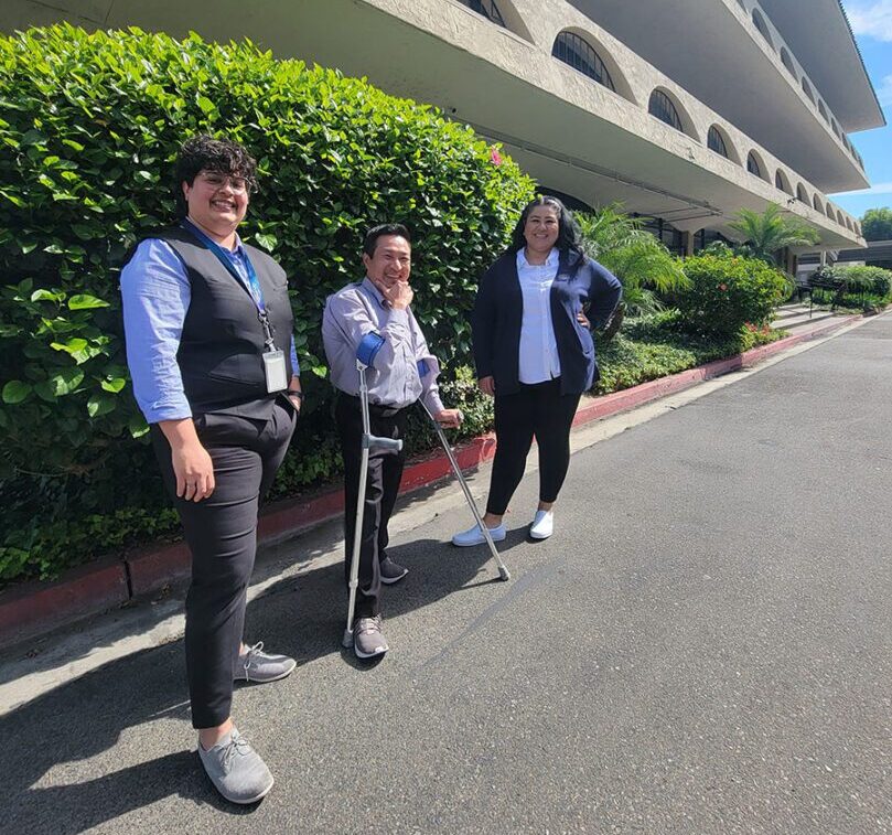 Pictured, DMC PAS team Michelle, Kevin and Judith standing outside of DMC smiling. Michelle and Judith have one hand on hip and Kevin has hand on chin, thinking.