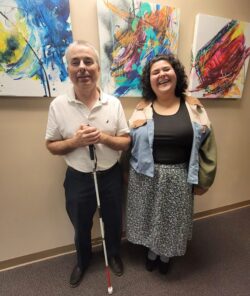 Pictured Independent Living Skills Team left -Paul standing with mobility cane and right -Anita standing inside DMC in front of an art wall of paintings.