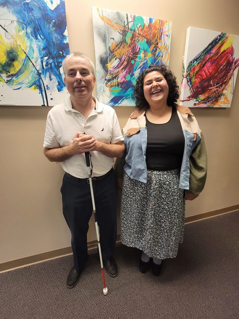 Pictured Independent Living Skills Team left -Paul standing with mobility cane and right -Anita standing inside DMC in front of an art wall of paintings.