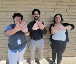 Standing in front of a tan wall is the Community Transitions Team Michelle, Babar and Christabel signing in ASL “Community” holding hands flat using the other hand pressing across the other fingers.