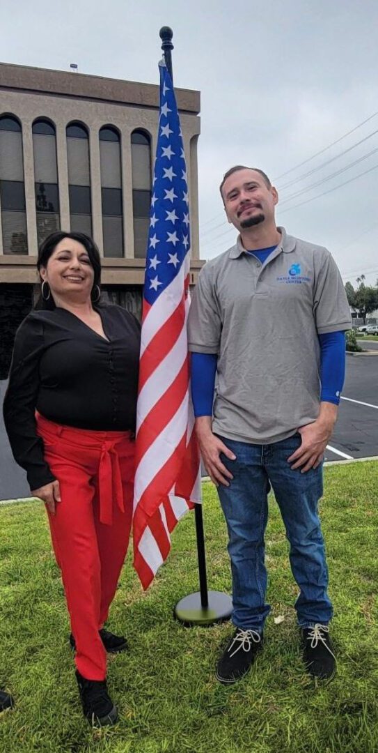 DMC’s Marisol, Veterans Independence Program Advisor and right – Fernando, Community Living Advocate standing proudly representing the VIP Services Team.  They stand united together with the U.S. Flag in the center outside of DMC offices on the grass. 