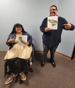 : DMC’s DMC Youth Connect Team smiling and pointing coast-to-coast directives in front of a light blue wall. Left: Nelly Gomez, Lead Youth and Information Coordinator holding a picture of California State in green with white lettering and Right: Alan Cruz, Youth Community Liaison and California State Independent Living Council holding a picture of South Carolina in green with white lettering.