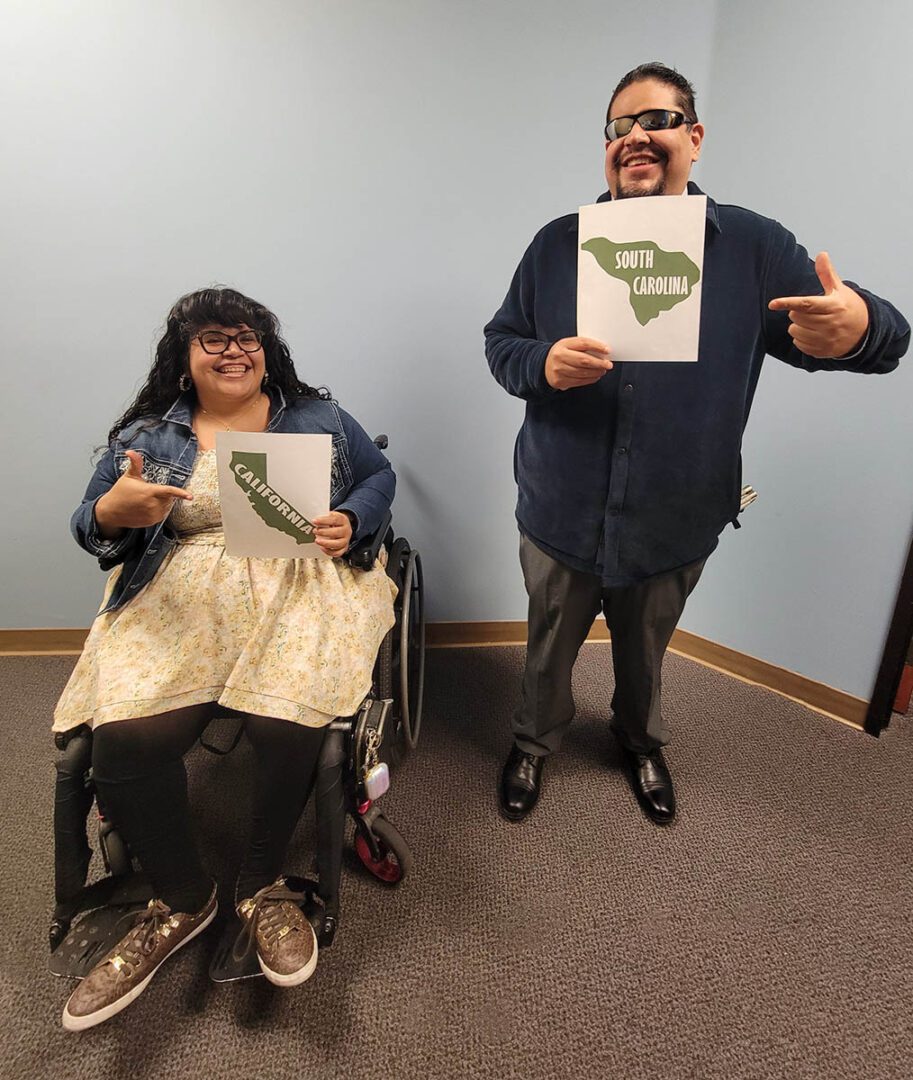 : DMC’s DMC Youth Connect Team smiling and pointing coast-to-coast directives in front of a light blue wall.  Left: Nelly Gomez, Lead Youth and Information Coordinator holding a picture of California State in green with white lettering and Right: Alan Cruz, Youth Community Liaison and California State Independent Living Council holding a picture of South Carolina in green with white lettering. 