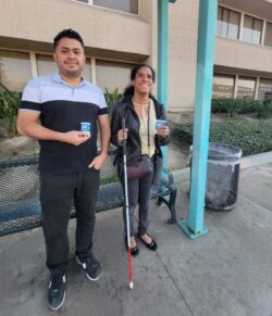 Pictured: DMC’s Lead Home Access Specialist and MMPP Trainer with DMC’s ILS Instructor for the Blind and OC Access user, Danyelle Cerillo at Bus Stop each standing in front of the bench smiling and each holding an OC Bus Day pass.