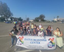 Photo of Team DMC at Pride Parade 2024 all standing outside in a parking lot with the Dayle McIntosh Center banner. The Banner is being held by six people and features the DMC logo in rainbow colors on each side of the banner.
