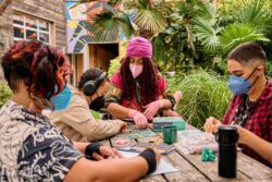 Four masked disabled people of color sit at a table outdoors, immersed in arts and crafts. Two people are working on a puzzle, one wearing arthritis gloves and the other with noise-canceling headphones. One person is shaping modeling clay, and one person is filling in a coloring book while wearing wrist support wraps. Two stim fidgets rest on the table, and a portable air purifier can be seen in the foreground to the right.