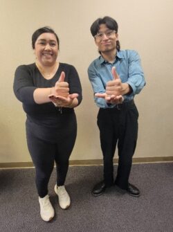 Pictured: Standing in front of beige wall at DMC, Information and Referral’s Bianca (left) and Front Office Volunteer Johan (right) both doing ASL sign for “Help” with thumbs up sitting in palm of hand.