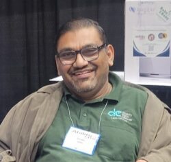 Pictured Bhumit Shah, DMC Board of Directors Member At-Large, wearing glasses, a green shirt and smiling.