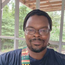 Pictured: Griffin Dooley wearing glasses with mustache sitting outside. He is wearing a grey shirt with a yellow, red and green colored strap on his shoulder.