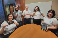 : Pictured – DMC Blind/Low-Vision Services Team behind a round table- everyone is smiling holding a special Assistive Technology device inside of DMC Team member are wearing DMC company T-shirts.