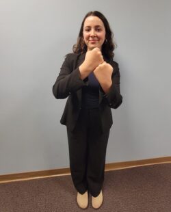 DMC’s Systems Change Advocate, BreeAnna Adner wearing black suit smiling as she does ASL with her hands signing ‘advocacy’ in support of her work at DMC.