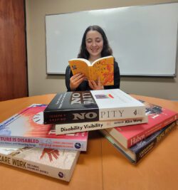 Pictured DMC Systems Change Advocate BreeAnna at DMC table filled with advocacy books smiling as she reads with a white dry erase board behind her.