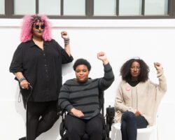 Torso level photo of three Black and disabled folx (a non-binary person holding a cane, a non-binary person in a power wheelchair, and a femme on a folding chair) raising their fists on the sidewalk in front of a white wall.