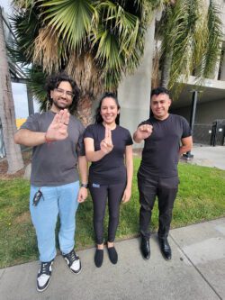 PICTURED DMC’s MMPP Team signing B-U-S in ASL standing on sidewalk with palm fronds behind them.