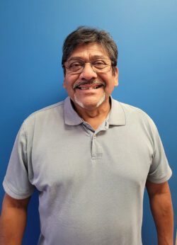 Pictured Board Vice President, David Flores wearing glasses and grey shirt smiling in front of blue wall