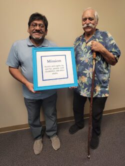 DMC’s Board Vice President David Flores with Board President, Tony Clement – both smiling proudly holding DMC’s Mission Statement in a blue frame which reads “Access and equity by, and for, people with disabilities and older adults."