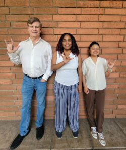 Pictured above: Deaf Services Team Jet, Pamela and Doryna all signing “I Love You” (ILY) in ASL standing together outside in front of a brick wall smiling.