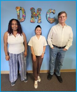 Pictured above: Deaf Services Team Pamela, Doryna and Jet standing inside DMC offices in front of blue wall with DMC colorful mosaic wall display of letters