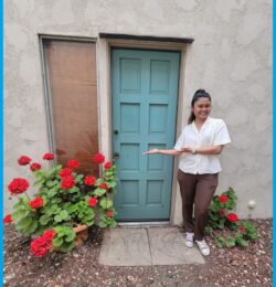 Pictured above: Doryna, Deaf Housing Coordinator, standing in front of teal door surrounded by red geranium bushes with slate slab walkway porch saying “Welcome Home.”