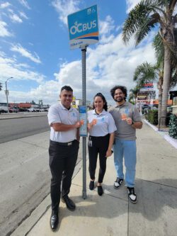Pictured MMPP Team holding bus passes at the bus stop smiling as they stand on the sidewalk.