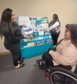 DMC Front office team at teal colored flyer kiosk. Bianca is showing Nelly a new flyer. Paola is organizing groups of flyers at the kiosk.