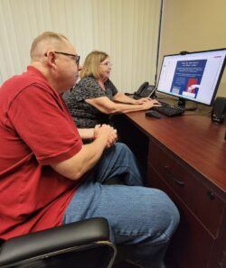 Two older adults sitting at desk looking at computer monitor ready to share DMC’s