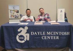 DMC's PAS Recruitment Specialist Kevin Dinh and Community Living Advocate Fernando Olivarez at 2025 CSUN Conference at DMC's booth with resource information materials on the table and DMC's blue table cloth with logo in white featured representing DMC