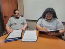 Image Description: DMC Youth Connect Team Alan and Nelly smiling wearing DMC staff shirts sitting at table going over program plans