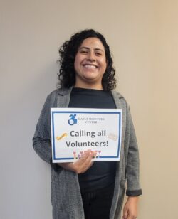 Calling all volunteers with DMC logo sign being held by Volunteer Daysi smiling inside the office at DMC