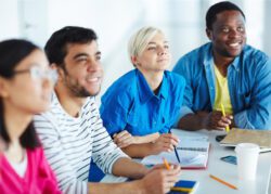 A diverse group of four adults sit at a table smiling and attentively looking forward. They have notebooks and pens in front of them, participating in a training.