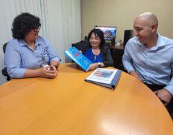 Pictured DMC VIP Team Michelle, Marysol and Eric gathered together around a table with Marysol sharing the VIP materials packet inside DMC