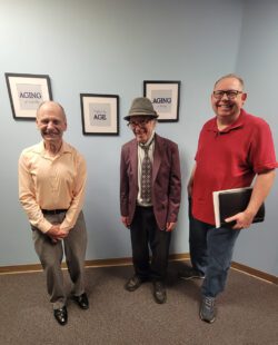 Pictured three older adults smiling standing together in front of artwork on a blue wall which reads “Aging, Age, Aging” to celebrate transitional milestones.