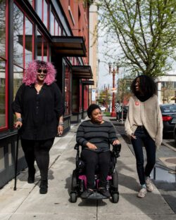 Three Black and disabled folx smile and stroll down a sidewalk side by side. On the left, a non-binary person walks with a cane in one hand and a tangle stim toy in the other. In the middle, a non-binary person rolls along in their power wheelchair. On the right, a femme gestures mid-walk.