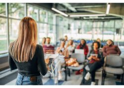 A person giving a presentation in front of a diverse audience. The seating area is blurred, making the faces of the group indistinguishable.