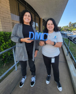 Volunteer Daysi standing with Volunteer Coordinator Raquel on ramp outside holding blue DMC sign