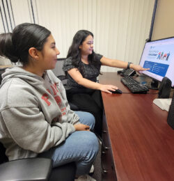 Pictured DMC Volunteer Coordinator Raquel showing Volunteer Sam the Volunteer flyer on the website at a desk inside DMC.