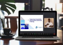 A laptop on a wooden table next to a coffee cup, displaying a virtual training titled “Improving Accessible and Inclusive Services Training” with a sign language interpreter visible on the screen. The logo of the Dayle McIntosh Center is displayed beneath the title.