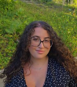 Gracie Doran, Board Member At-Large smiling with long curly brown hair and glasses, wearing a floral top with grass in the background outside