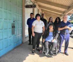 DMC Directors and Leads together outside on sunny day next to set of double blue doors. L-R: Front Row: Michelle, Nelly, Paul, Back row Ivan, Jarvous, Brittany and Pamela.