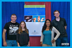 DMC Deaf Services Team Jet and Pamela at Deaf Nation Expo standing in front of blue and red curtains in front of ASL poster smiling