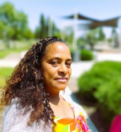 Grace Yhap with long curly hair looking at camera from the side. She is wearing bright floral top with neighborhood and green shrubbery in the background.