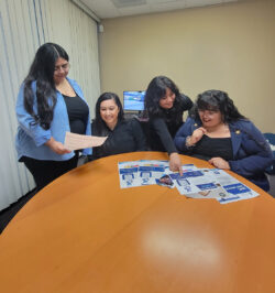 DMC I&R team at a desk. Mareli is showing Nelly a new flyer. Paola and Bianca are looking at another flyer.