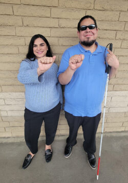 DMC AT Coordinators, Lizette Martinez and Alan Cruz smiling wearing blue tops and black pants standing outside in front of beige brick wall signing “AT” in ASL Finger Spelling.
