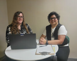 DMC Directors Brittany and Michelle sitting at table with computer and stack of papers inside DMC discussing plans smiling.