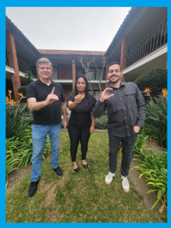 Pictured above: Deaf Services Team Jet, Pamela and Tobias signing D-M-C in ASL standing outside in courtyard with Bird of Paradise orange foliage behind them