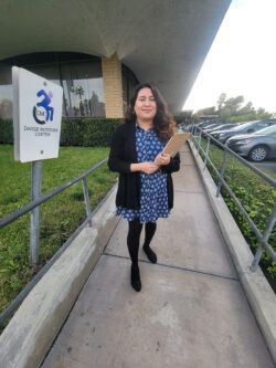 Transitions Coordinator, Lizeth standing on ramp with clipboard outside DMC next to DMC blue logo sign with arrow pointing toward the door